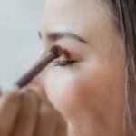 Custom Makeup - Side view of crop young female customer with ling brown hair and sitting in light studio closed eyes while anonymous makeup artist applying eyeshadows