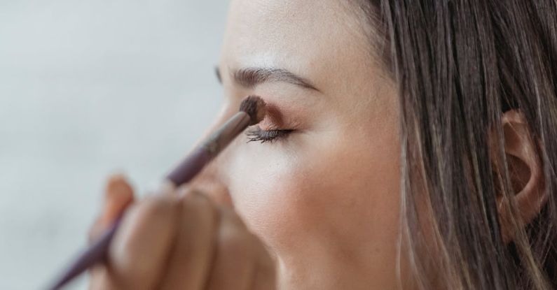 Custom Makeup - Side view of crop young female customer with ling brown hair and sitting in light studio closed eyes while anonymous makeup artist applying eyeshadows