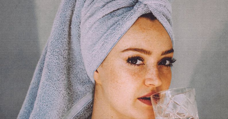 Healthy Hair Ends - Young woman with hair wrapped with towel and bared shoulders drinking water and looking at camera