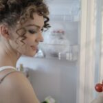 Healthy Food Hair - Side view of content female in casual wear standing in kitchen near opened fridge and picking fruit for breakfast