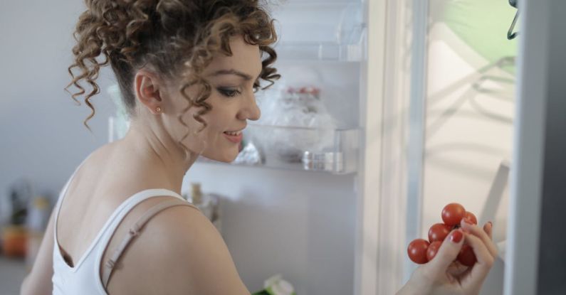 Healthy Food Hair - Side view of content female in casual wear standing in kitchen near opened fridge and picking fruit for breakfast