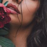 Organic Makeup - Dreamy female with black hair wearing white blouse with bare shoulder looking at camera while standing near wall with red flower in hand