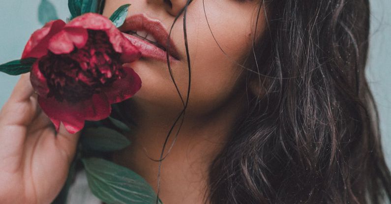 Organic Makeup - Dreamy female with black hair wearing white blouse with bare shoulder looking at camera while standing near wall with red flower in hand