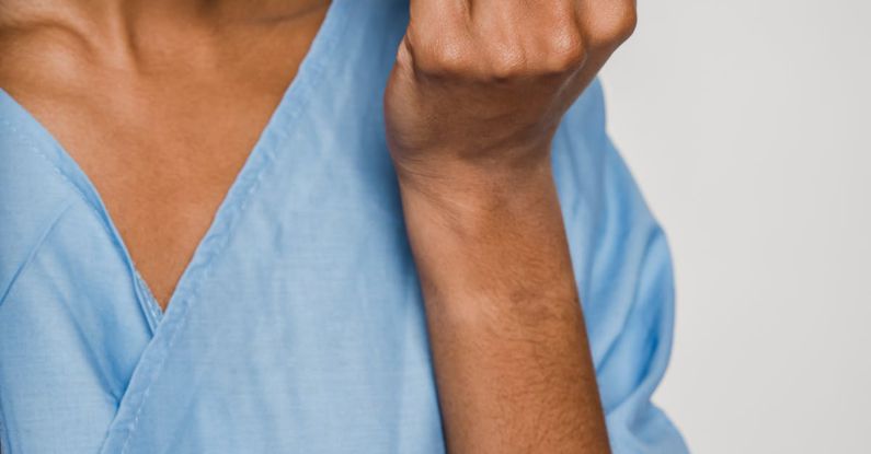 Surgery Risks - Unrecognizable African American female nurse in blue uniform putting on protective mask while standing against white wall